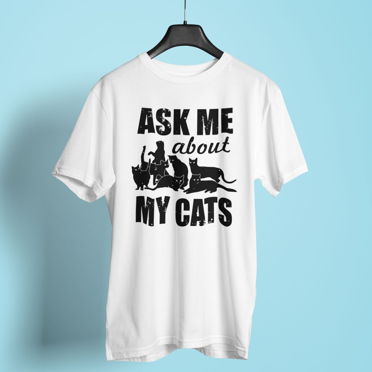 White t-shirt with 'Ask Me About My Cats' text and cat illustrations on a hanger against a light blue background