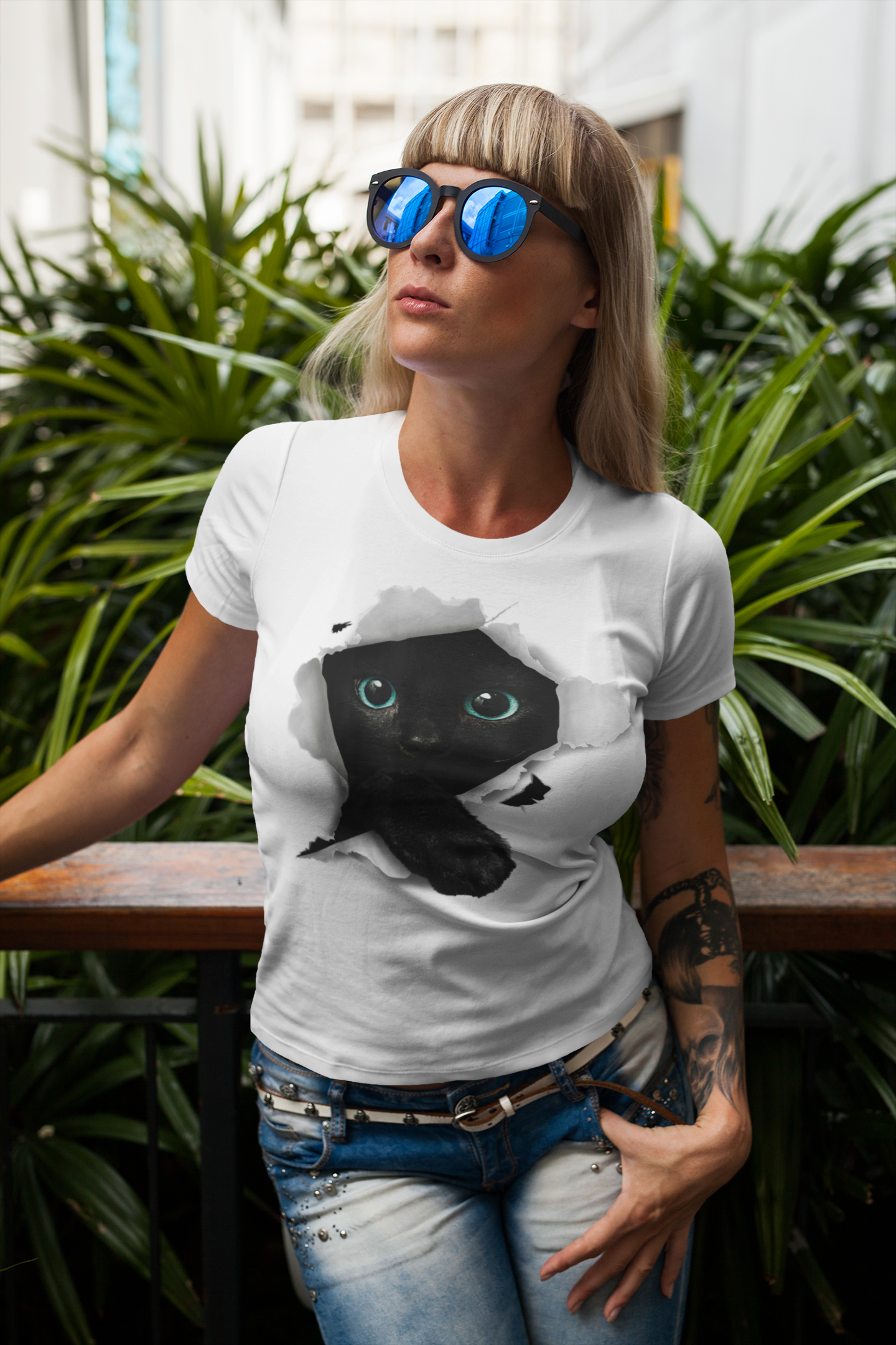Woman wearing a white t-shirt with a black cat graphic, standing outdoors with plants in the background.