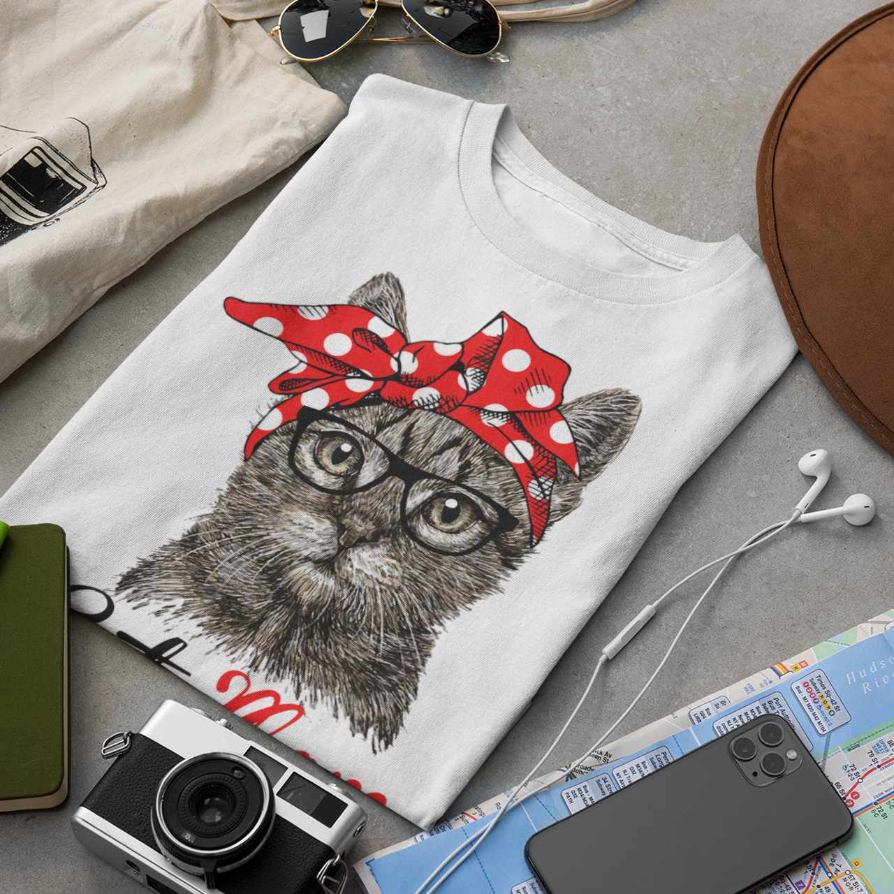 T-shirt with a cat design wearing a red bandana, surrounded by a camera, hat, map, and other items on a gray surface.