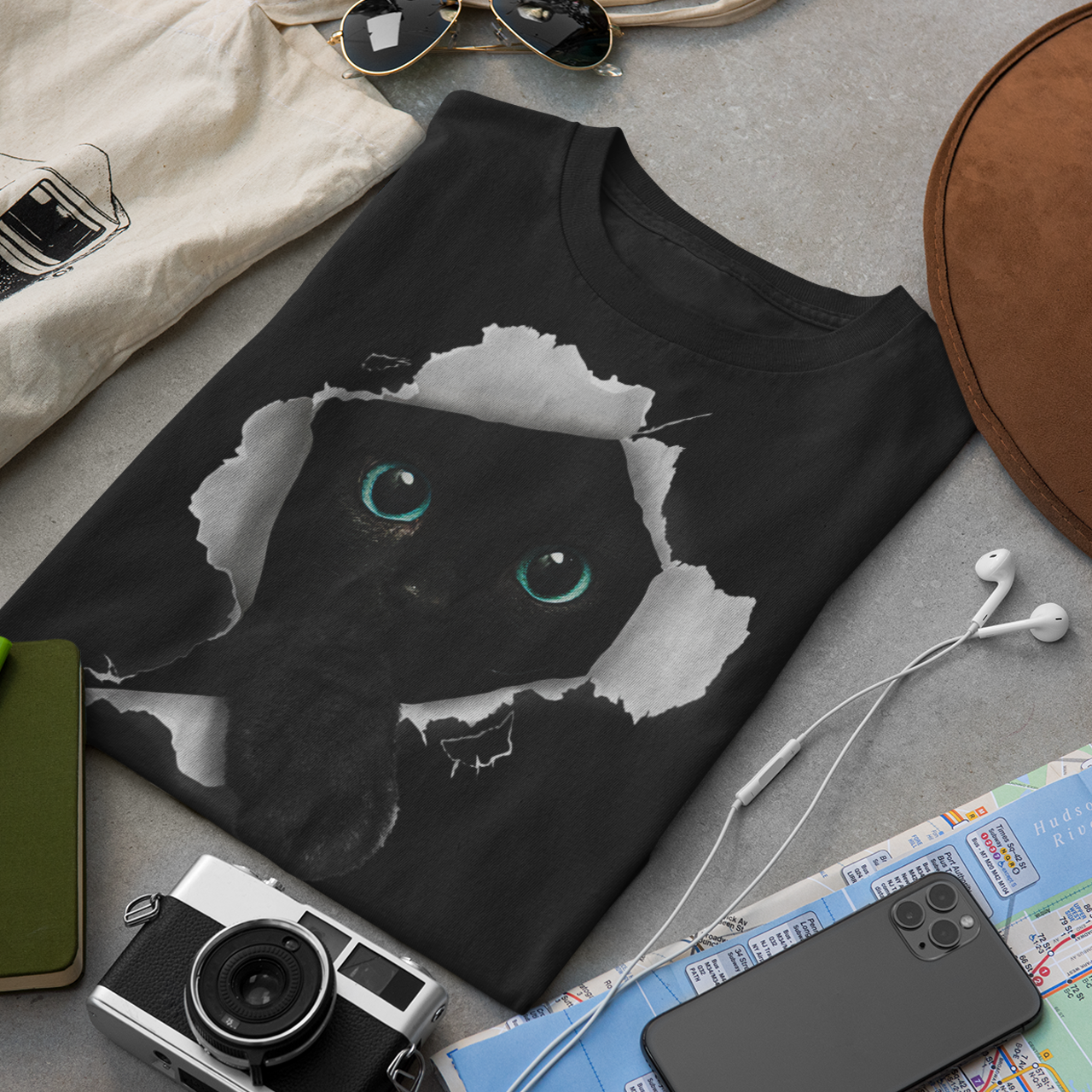 Black t-shirt with a cat design on a gray surface with accessories like a camera, hat, and map.