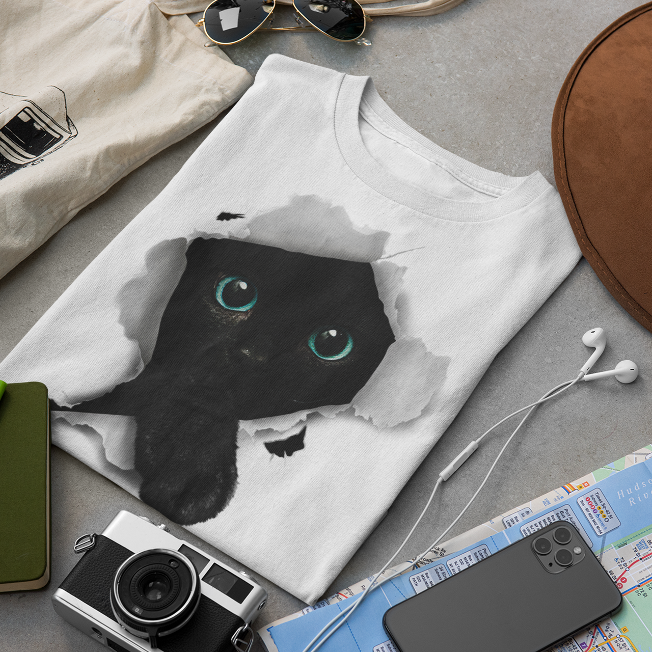 White t-shirt with black cat design, camera, hat, map, and phone on a gray surface